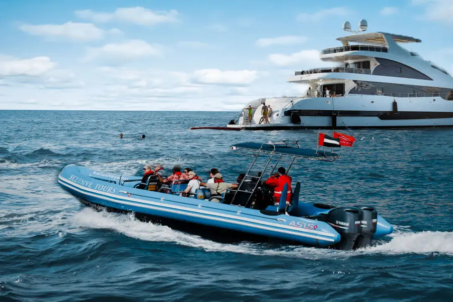 A blue inflatable boat with passengers cruises near a large yacht in open water.