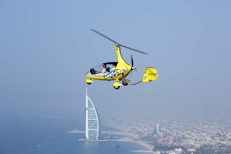 A gyrocopter flies over the city with the iconic Burj Al Arab visible in the background against a clear sky.