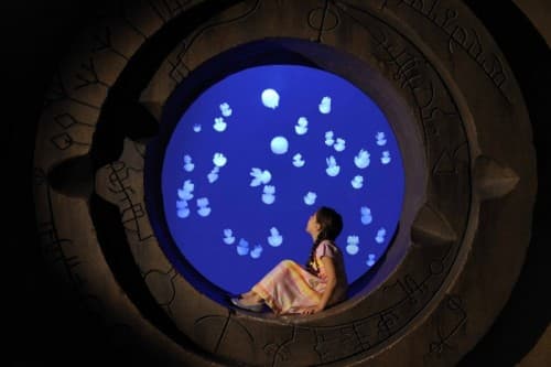 A girl gazes at floating bubbles while sitting in a round window during the Fish Tales Tour Kid Jelly Fish event.