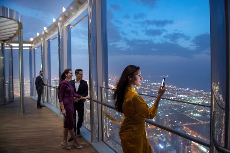 A woman in a yellow dress takes a photo with her phone on Burj Khalifa observation deck at twilight.