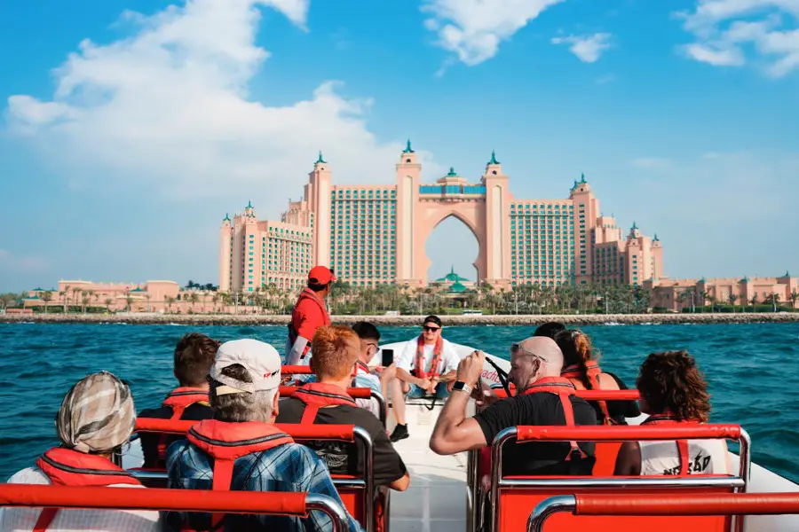 A group of people in life jackets sits on a boat, approaching a large, luxurious hotel in the background.