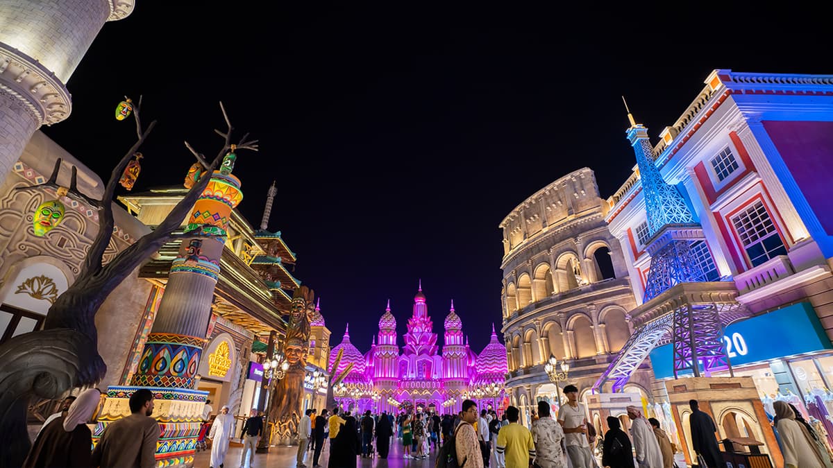 A group of people walking through Global Village Dubai at night, surrounded by shops and decorative lights.