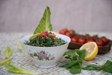 A decorative bowl of tabbouleh garnished with lettuce, mint, and lemon sits on woven mat.