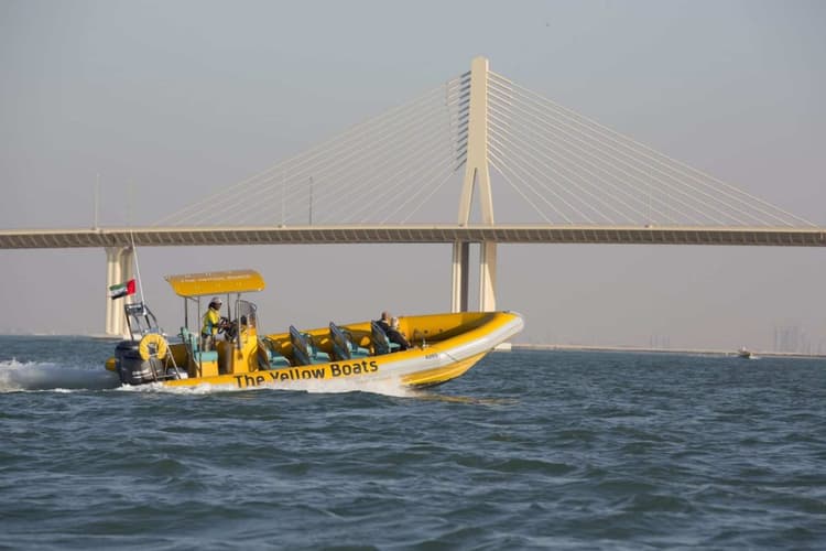 A yellow boat with people aboard floats in the water, set against the backdrop of Abu Dhabi City.