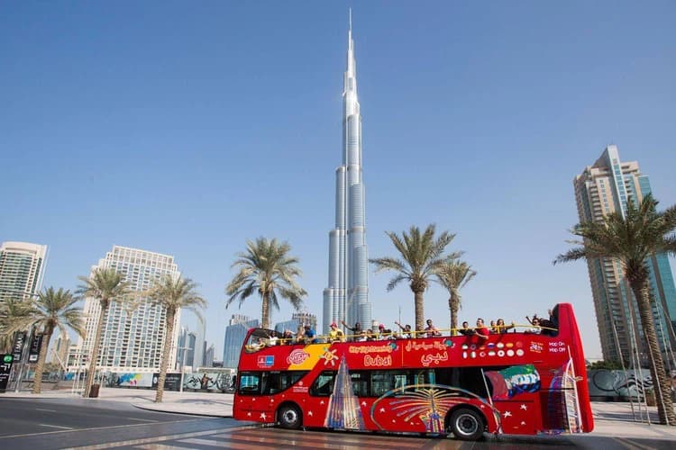 A vibrant red double-decker tour bus with tourists on top passes by palm trees