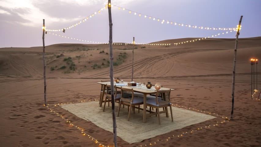 A serene desert dinner setup at sunset with a wooden table and chairs on a rug.