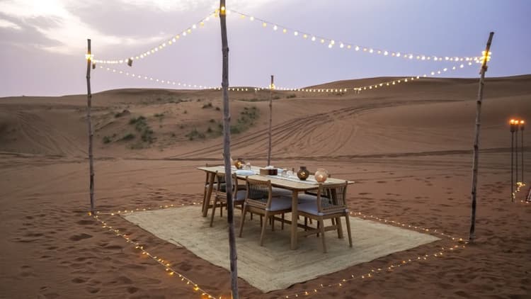 A serene desert dinner setup at sunset with a wooden table and chairs on a rug.