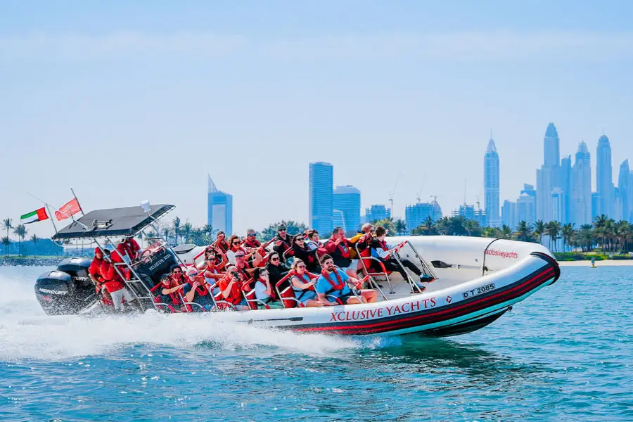A speedboat with people in red life jackets cruises through clear water, creating waves.