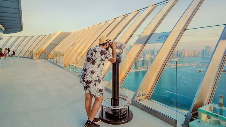 A man stands on the view at the palm, gazing out at a sprawling cityscape under a clear blue sky.