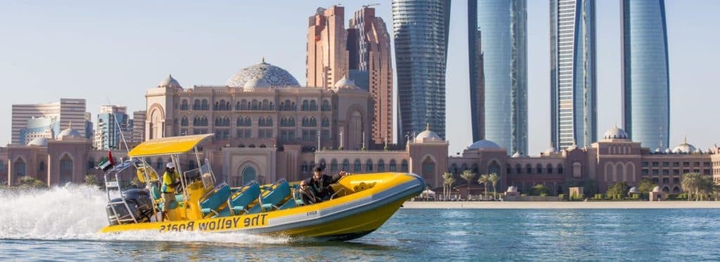 A yellow boat navigates the water with tall buildings of Abu Dhabi City in the background.