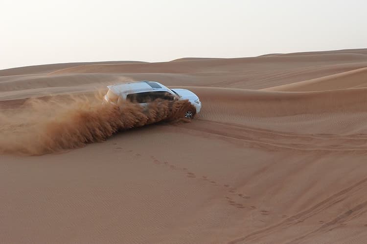 A white SUV navigates a sandy desert, kicking up a dramatic spray of sand.
