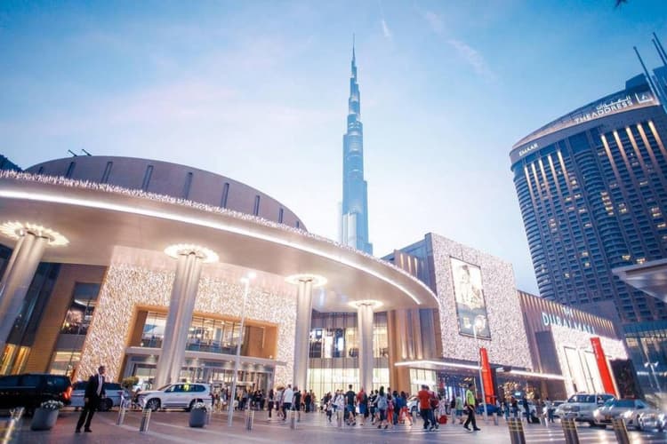 Busy shopping mall entrance at dusk with bright lights and digital screens.