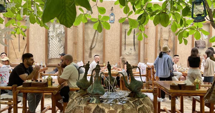A cozy outdoor café with leafy trees overhead, wooden benches, and a central stone fountain.