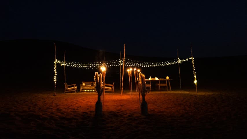 night scene in the desert features a lit seating area with a canopy of string lights and torches. Two wooden chairs and a table are warmly illuminated, set against a dark sky.