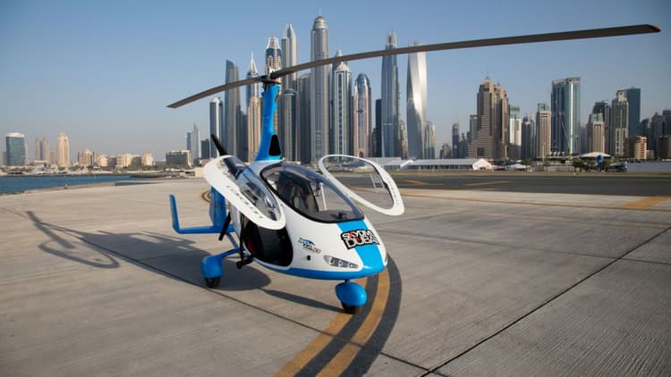 A gyrocopter on the tarmac with a city skyline in the background, labeled as the Gyrocopter Flight Helipad.