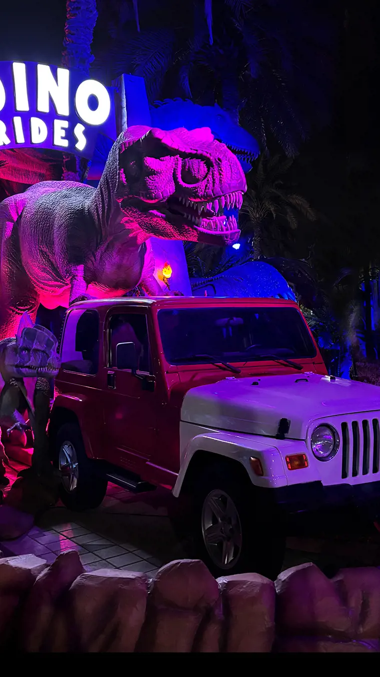 Large dinosaur model standing behind a jeep in Dinosaur Park creating a realistic prehistoric adventure scene.