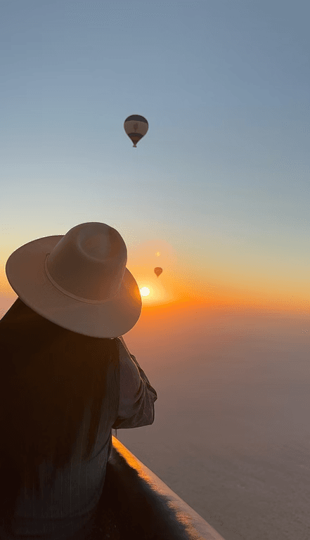 A person in a wide-brimmed hat admires a sunrise from a hot air balloon.