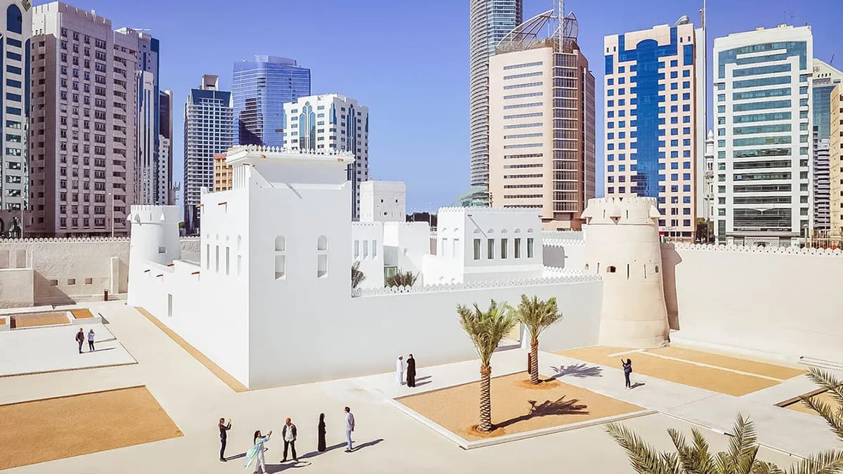 A white building with palm trees in front of tall skyscrapers against a clear blue sky. Qasr Al Hosn