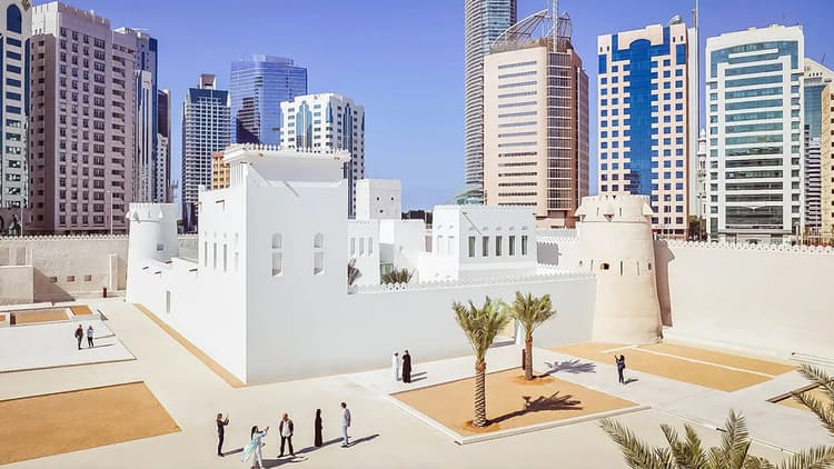 A white building with palm trees in front of tall skyscrapers against a clear blue sky. Qasr Al Hosn