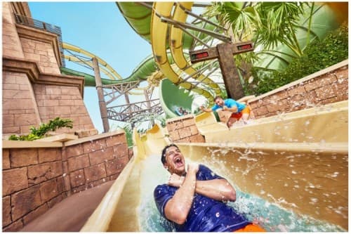 A man joyfully rides down a colorful water slide at a bustling water park on a sunny day.