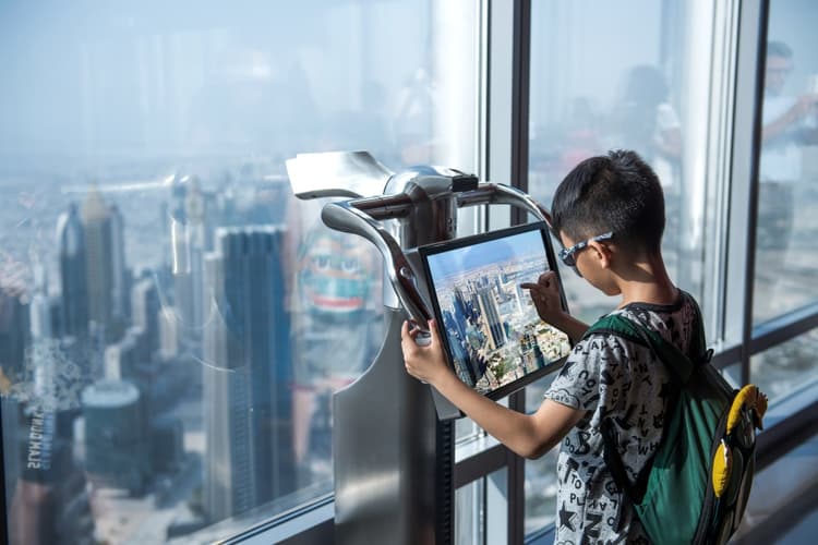 A young boy wearing sunglasses and a backpack interacts with a digital touchscreen displaying a cityscape from the Burj Khalifa's observation deck.