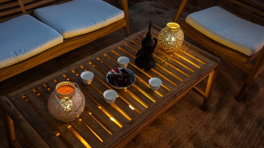 Cozy outdoor seating area with a wooden table, lanterns casting warm light, a teapot, cups, and a bowl of dates